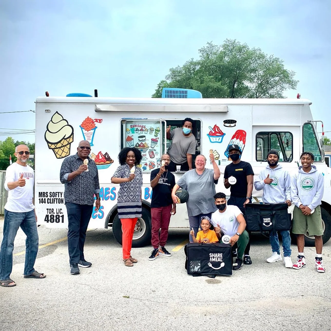 Swabir Shariff kneeling with a child in front of a crowd of youth, families and neighbours at an ice cream truck, Share-a-Meal bags in hand, at a Good Guides community event in Scarborough