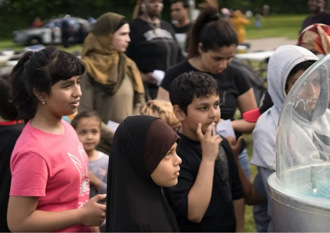 Children watching a cotton-candy machine spin at an outdoor community event, a girl in a pink shirt in the foreground